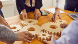 A set of wooden cogs on a table, with a group of businesspeople slotting them together.