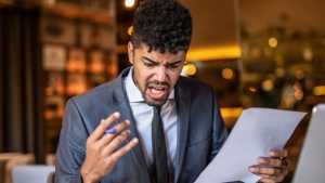 A young back male restaurant manager in a grey suit and tie, stood in front of the bar, holding a sheet of paper and pen, and grimacing.