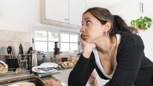 A youn woman with dark hair tied back in a pony tail, wearing a black jumper, with her chin in her hand, standing in a kitchen, wondering how to get food products to market.