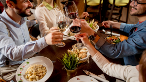 A restaurant table of four young businessmen toasting with glasses of red wine, with sharing platters of food on the table.