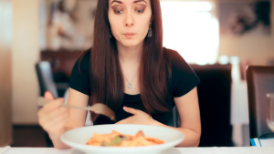 A woman with long brown hair, wearing a black t-shirt, holdind a fork, frowning, and picking at a plate of food.