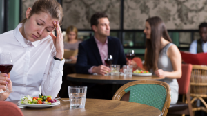 A young woman in a white short, sat on her own in a restaurant with a plate of food and a glass of read wine, holding her hand up to to her head, grimacing and looking down miserably.