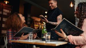 A young black male waiter, taking an order from two young black women holding menus in a restaurant.