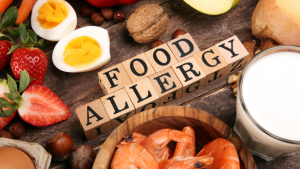 Wooden blocks spelling out FOOD ALLERGY surrounded by shelled prawns, mixed nuts, a glass of mike, half eggs, strawberries, and bread.