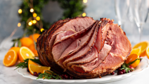 A glazed ham on a white platter, with red berries and orange slices, and a lit Christmas wreath in the background.