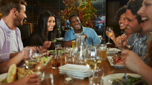 A diverse group of happy, smiling people eating a meal around a restaurant table filled with glasses and drinks.