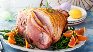 A whole glazed ham on a white plate, with orange slices and green beans, plus coloured Easter eggs in the background.