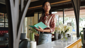 A female restaurant manager with long dark brown hair, holding a notebook, stood behind a bar.