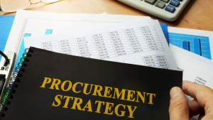 A black notebook with PROCUREMENT STRATEGY in large gold letters, on a desk with a pile of spreadsheets.