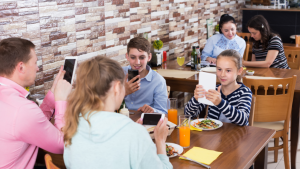 A young white mother, father, and Gen Alpha son and daughter, sitting at a restaurant table, ordering on tablets.