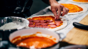 Hands making their own pizza with dough, passata and pepperoni at a restaurant pizza making experience.