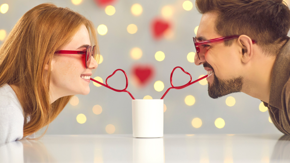 A young smiling couple wearing red heart-shaped sunglasses, sitting opposite each other over a white table, drinking from the same white drink with red heart-shaped straws.