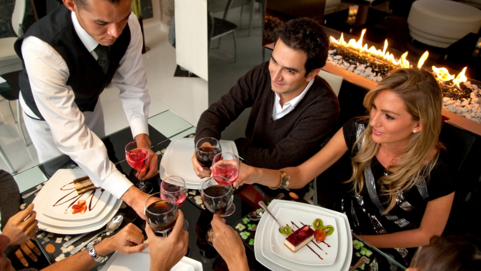 A table of fourr men and women having a toast with glasses of red wine as a waiter serves desserts on square white plates.