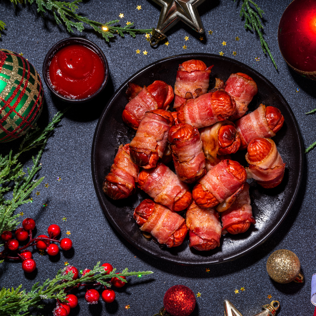 A black platter of pigs in blankets with a pot of tomato ketchup, Christmas red berries, greenery, and decorations.