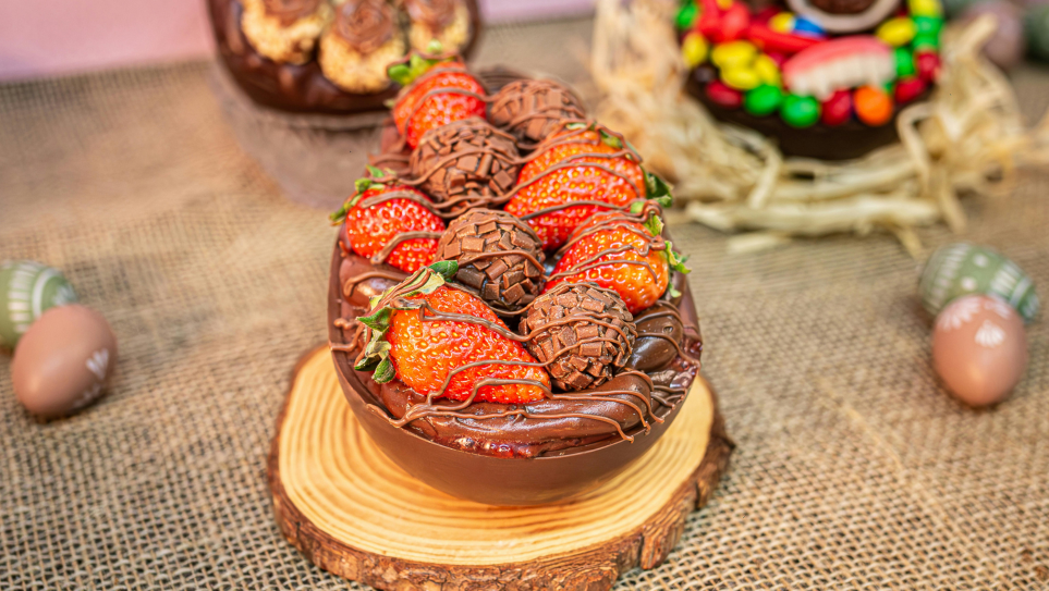 A half chocolate Easter egg filled with ganache, truffles and strawberries on a log slice, surrounded by painted eggs and desserts for Easter restaurant preparation.