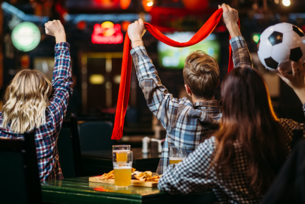 A table of football fans warching a match on a big screen, waving scarves and punching the air, with beers and food.