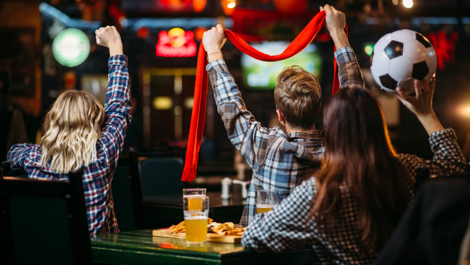 A table of football fans warching a match on a big screen, waving scarves and punching the air, with beers and food.