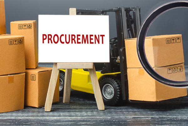 A white board on an easel wthe PROCUREMENT in red letters, in front of a forklift with piles of large boxes.