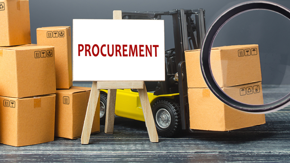 A white board on an easel wthe PROCUREMENT in red letters, in front of a forklift with piles of large boxes.